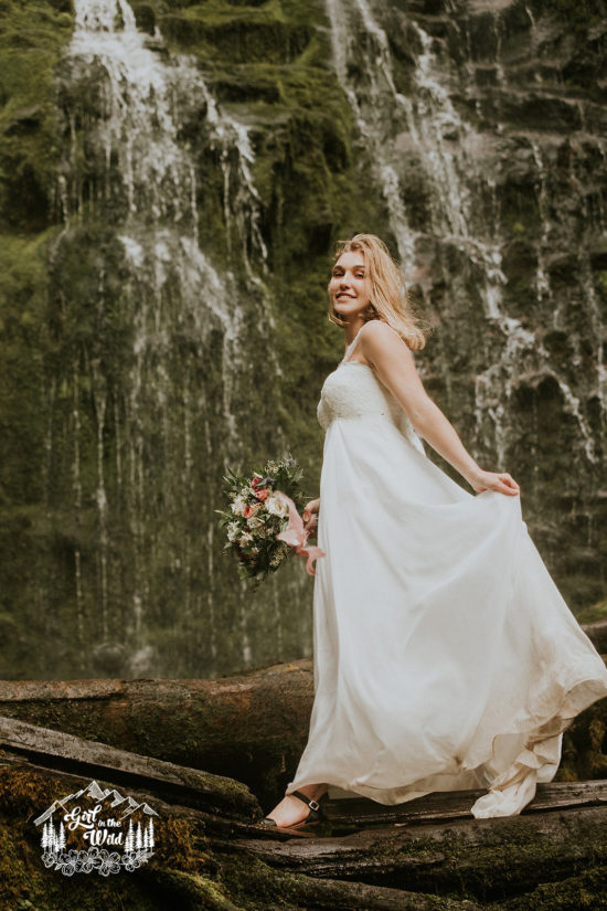 a bride steps carefully while holding her skirt up along a mossy log in front of a waterfall during her Sisters, OR Adventure Elopement