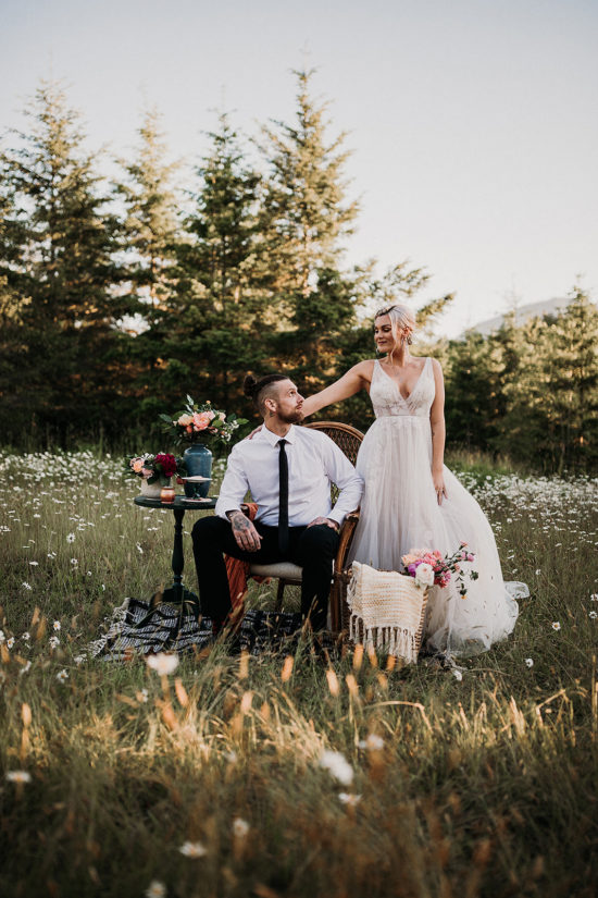 a groom in a suit sits in a chair and a bride in a tulle dress stands with her hand on his shoulder while they look at each other in the middle of a daisy meadow during their Olympic Peninsula Adventure Elopement